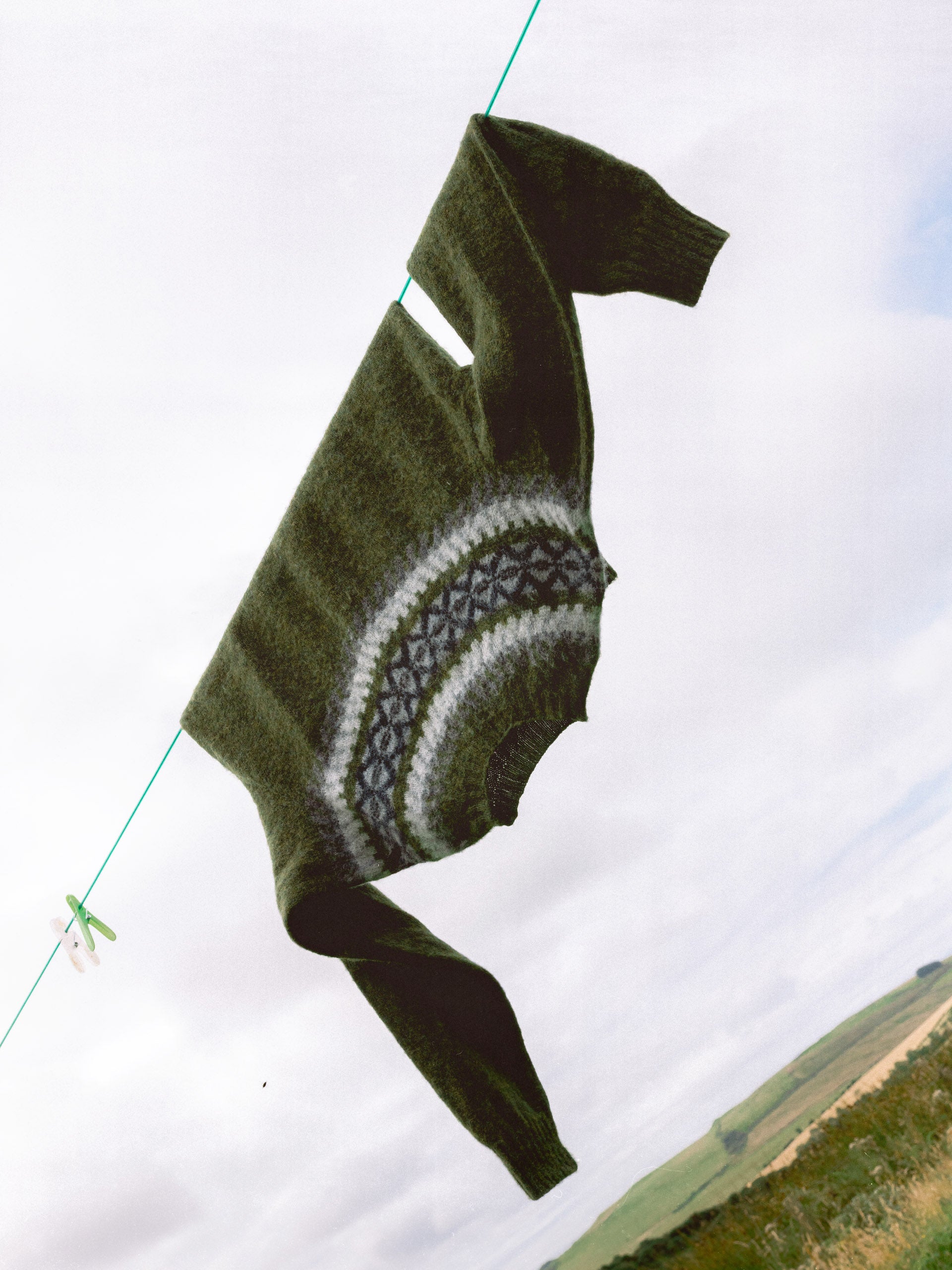 Olive knitted crew with a scottish traditional fairilse pattern hanging on a clothesline against a country background