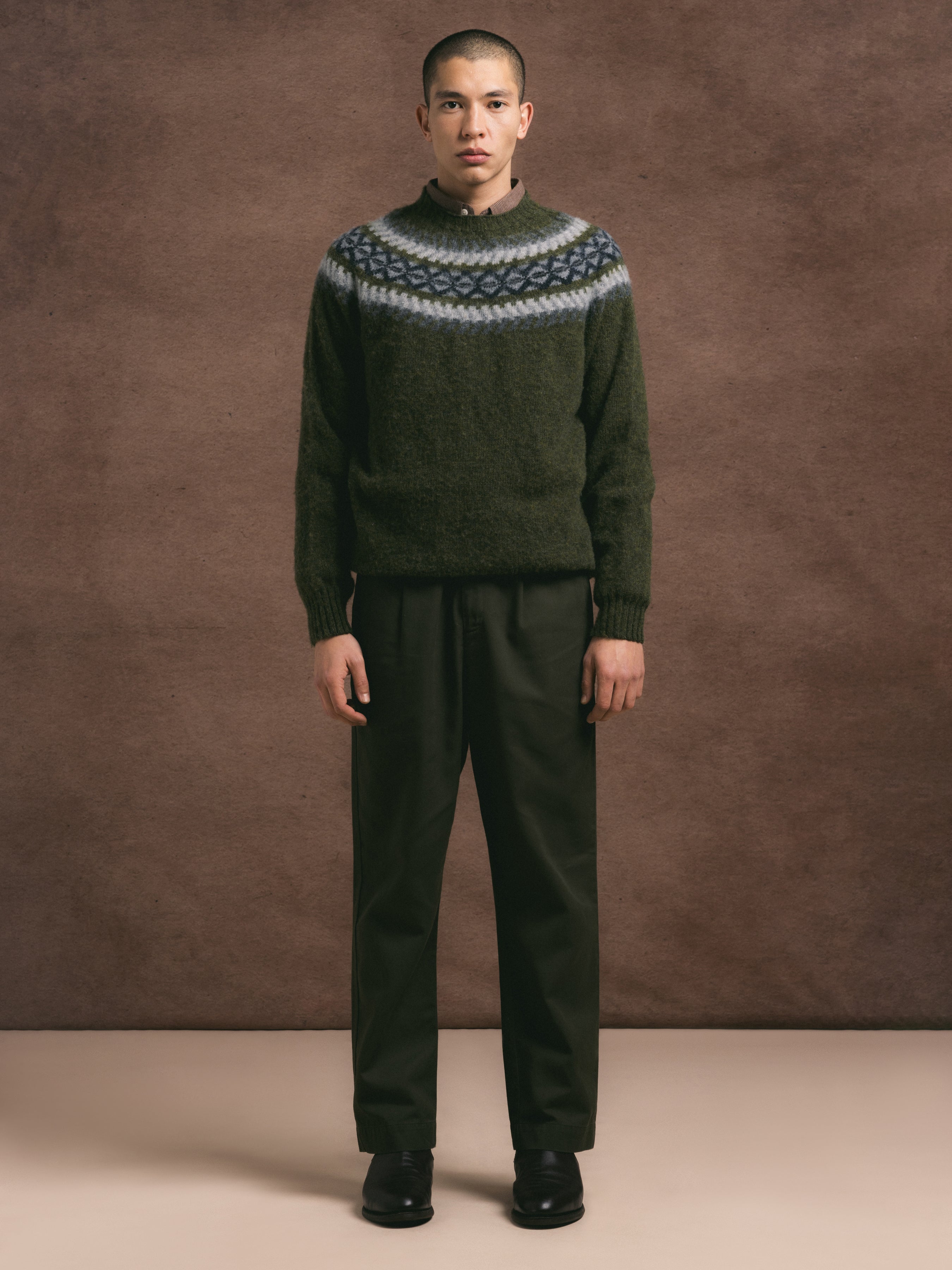 A model wearing a Scotish fairisle pattern olive crew over a shirt, with olive trousers against a textured background.