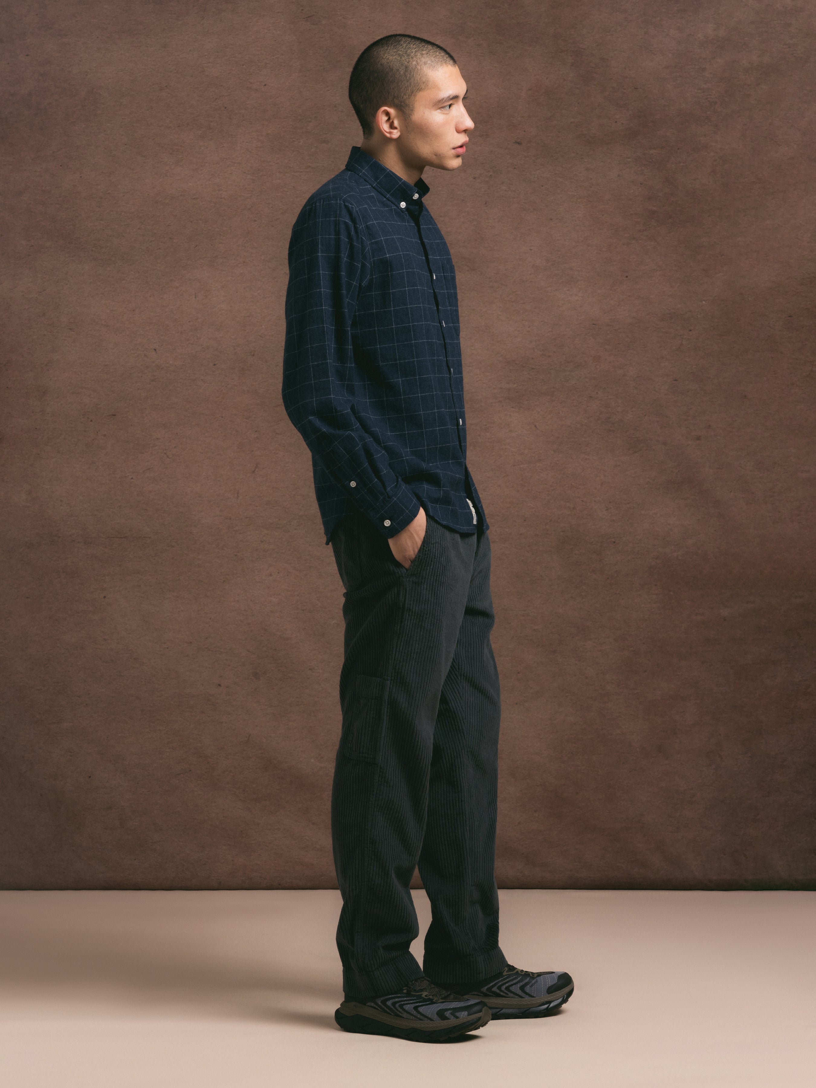 Man wearing a dark plaid shirt and grey pants against a brown background