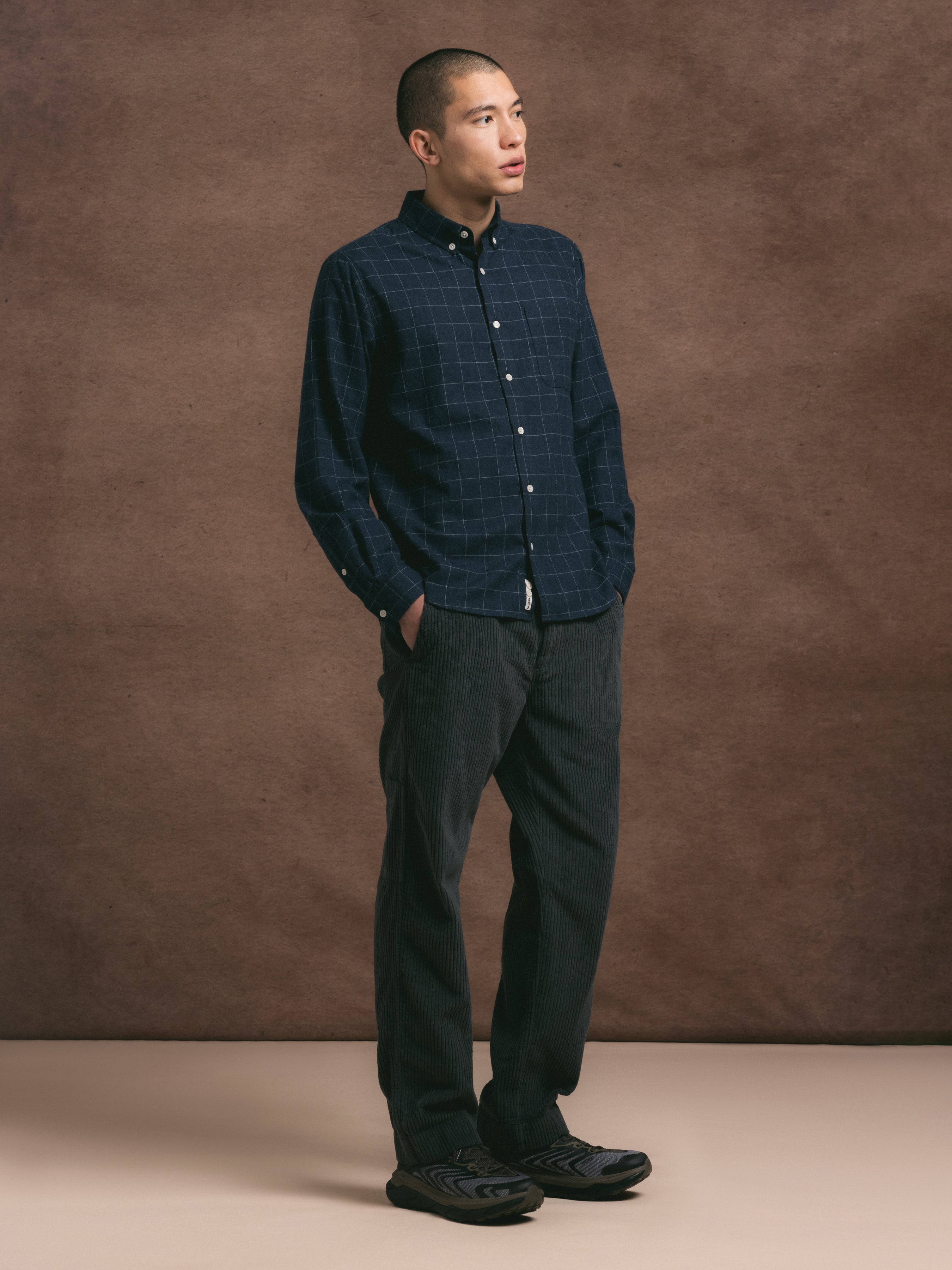 Man wearing a blue plaid shirt and dark grey pants against a brown background