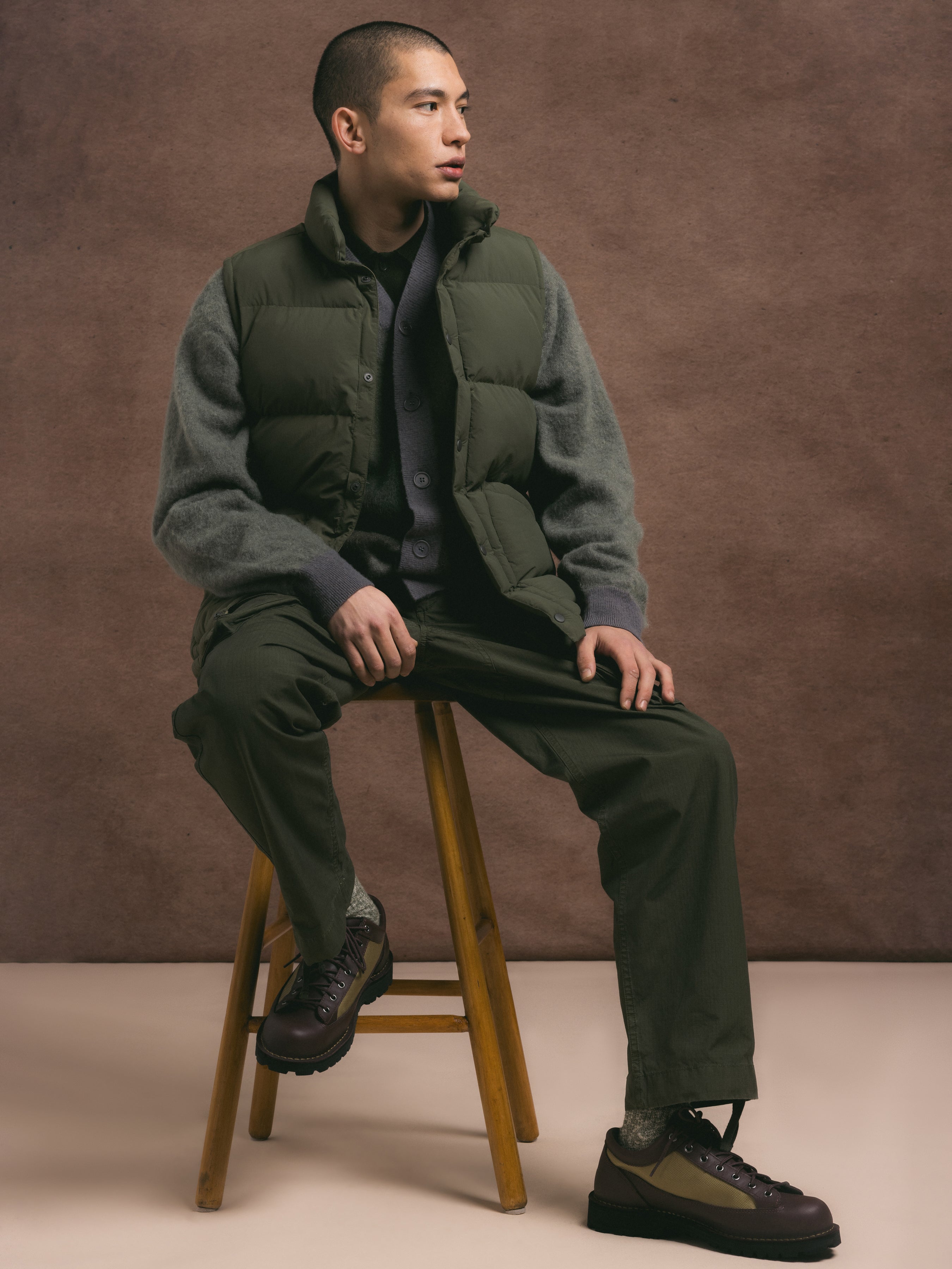 Man sitting on a wooden stool wearing a green puffer jacket and pants against a brown background