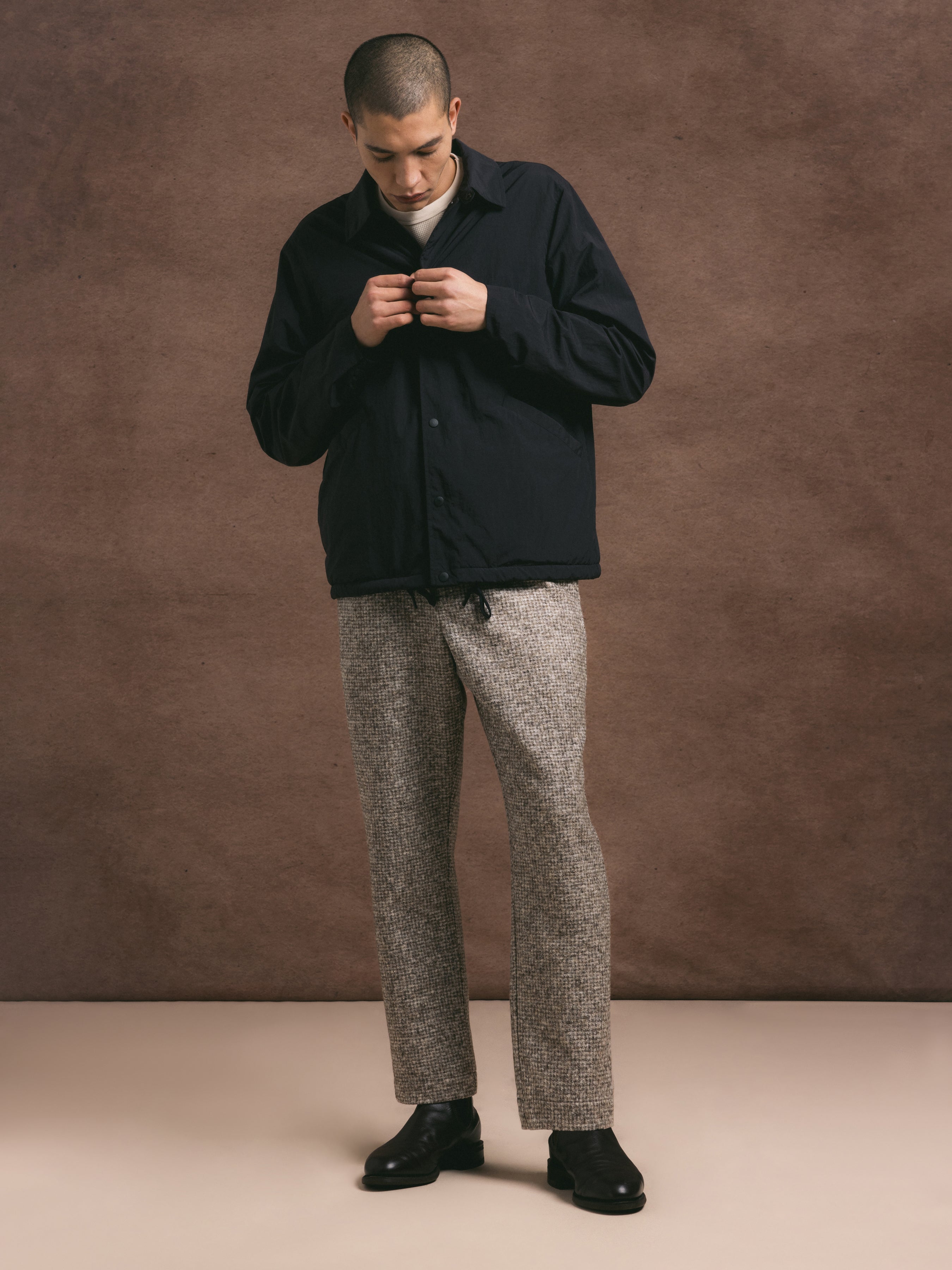 Man wearing a dark jacket and wool check taupe trousers, paired with black shoes, against a brown background
