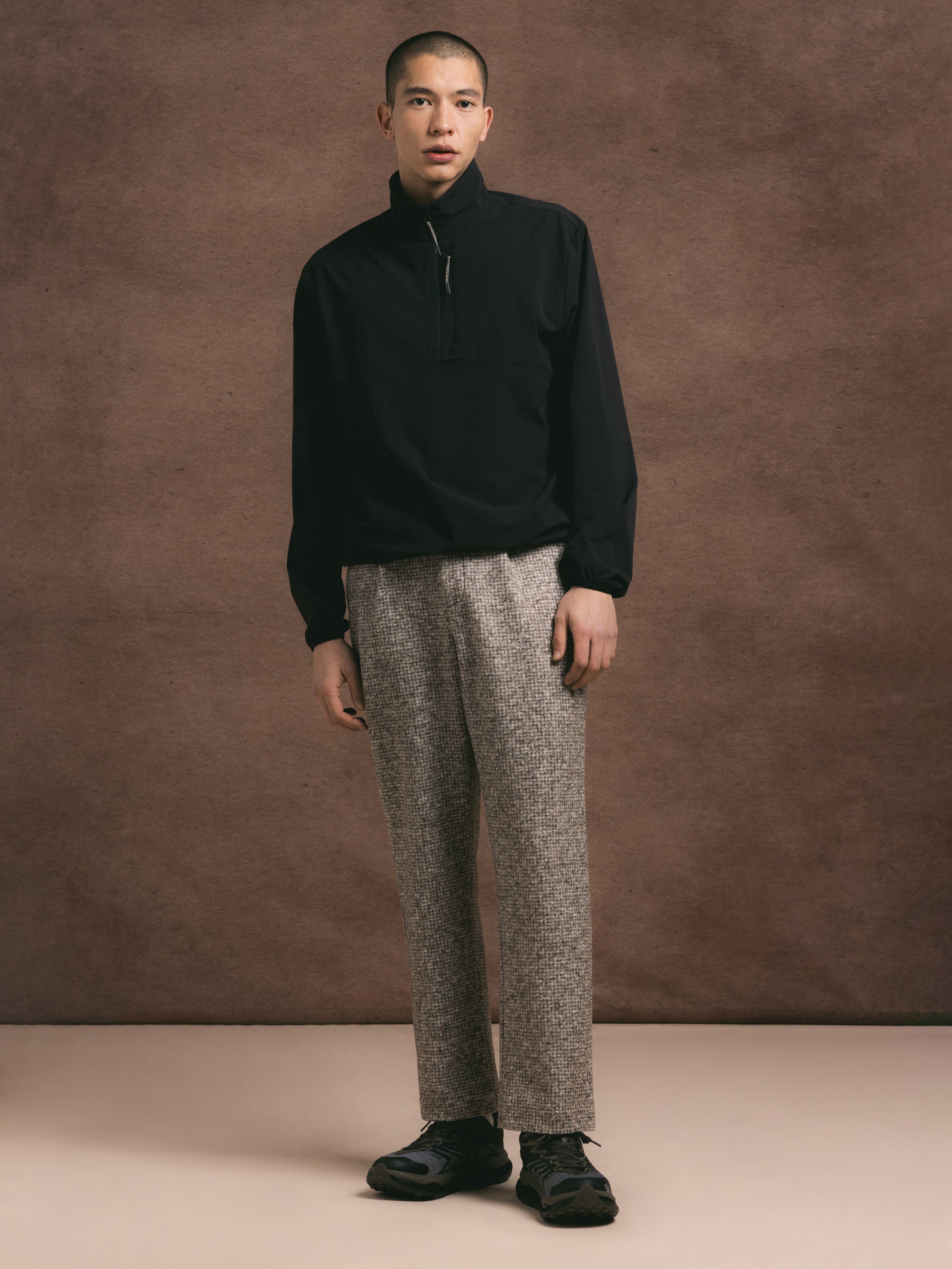 Man wearing a black quarter zip fleece and wool check trousers against a brown textured background