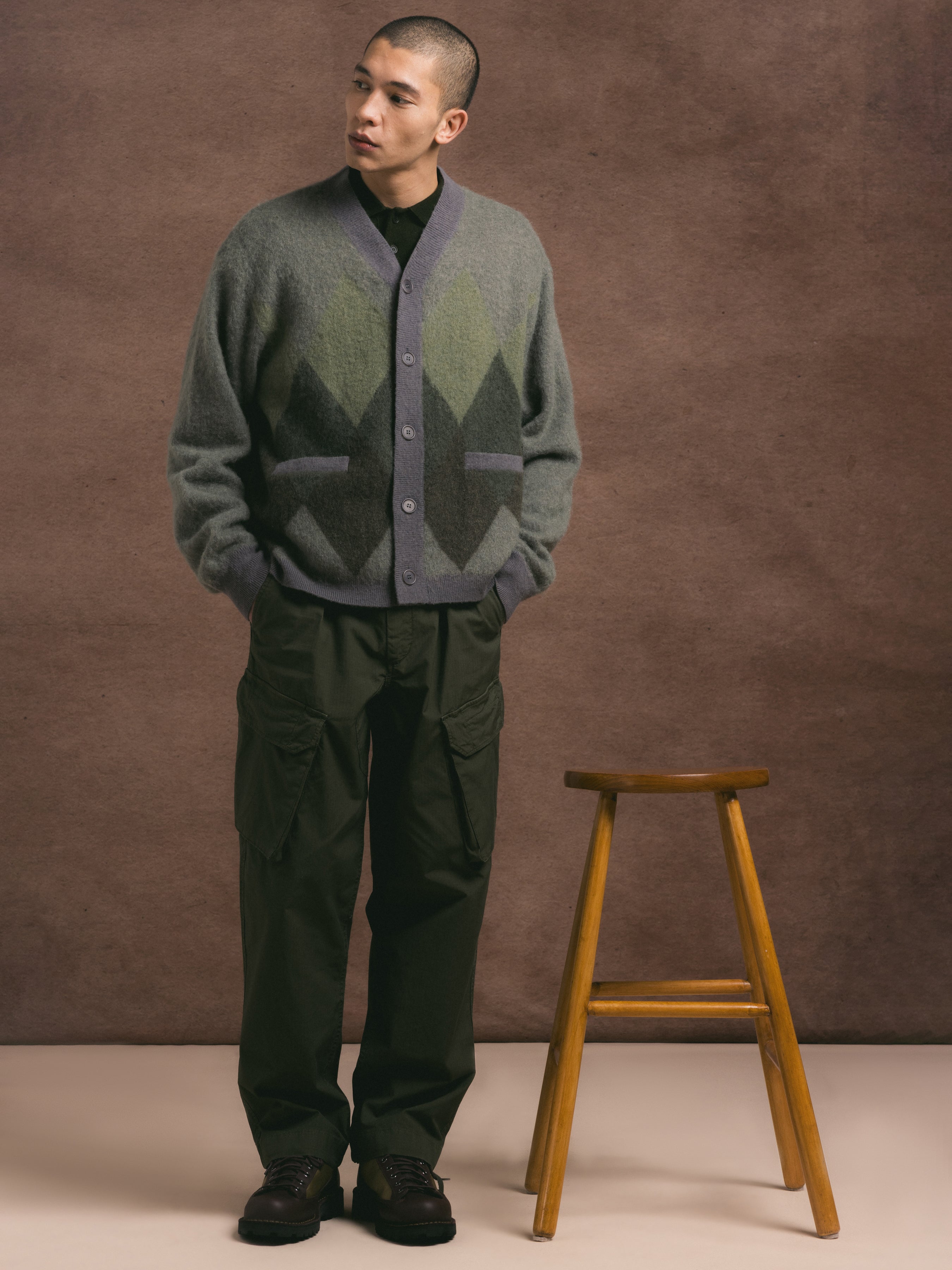 Man wearing a patterned cardigan and dark pants standing next to a wooden stool against a brown background