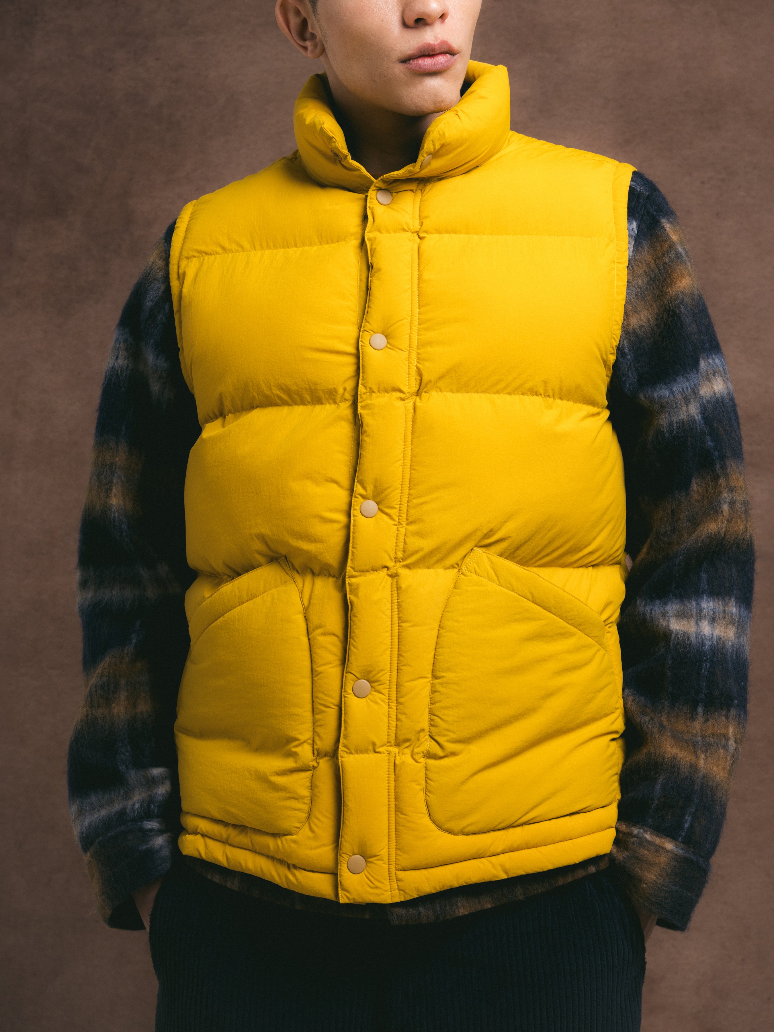 Person wearing a yellow puffer vest over a plaid jacket on a brown background