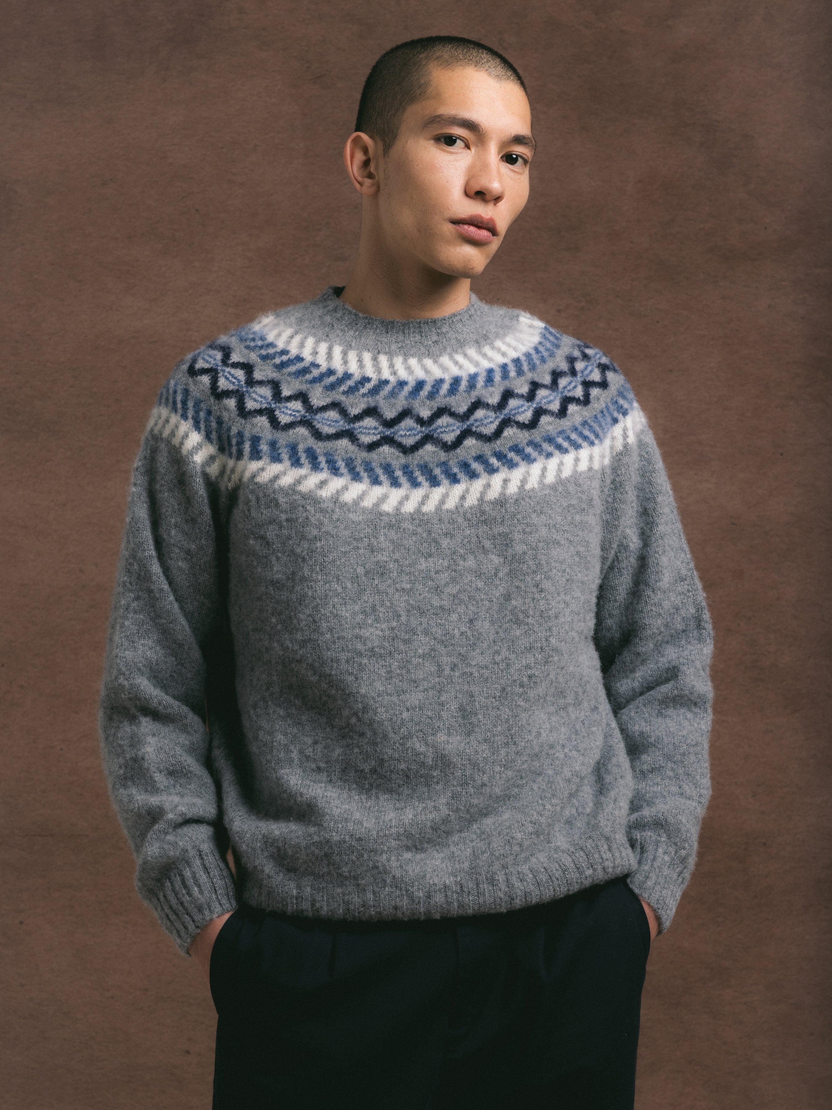 A model wearing a grey knitted scottish crew with a traditional fairisle pattern design, paired with dark trousers against a brown textured background