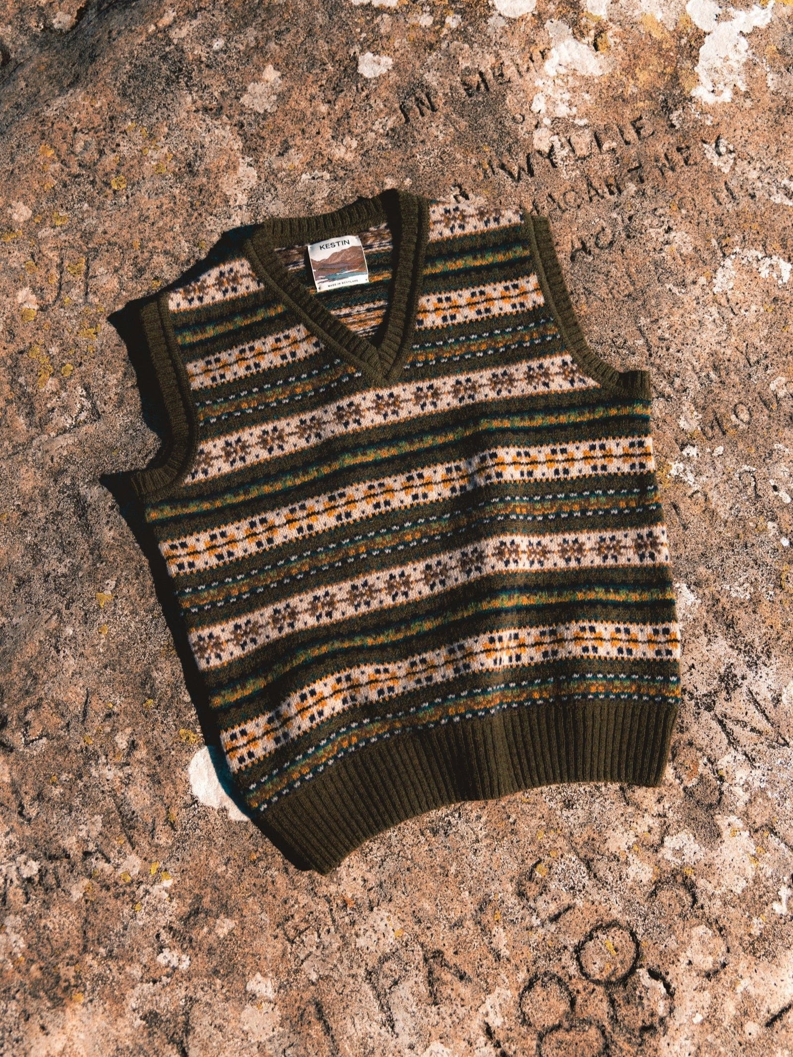 A Fairisle Sweater Vest in olive green, lying on a rock in Scotland.