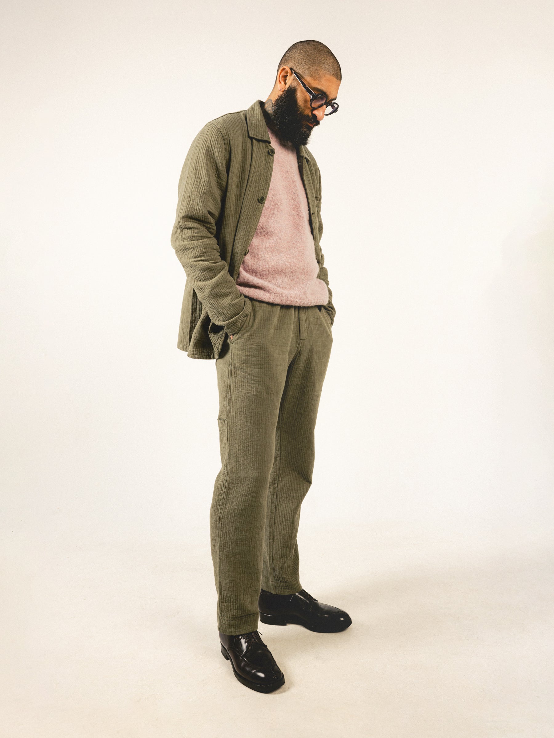 A man wearing a green chore coat and trousers with a pink sweater.
