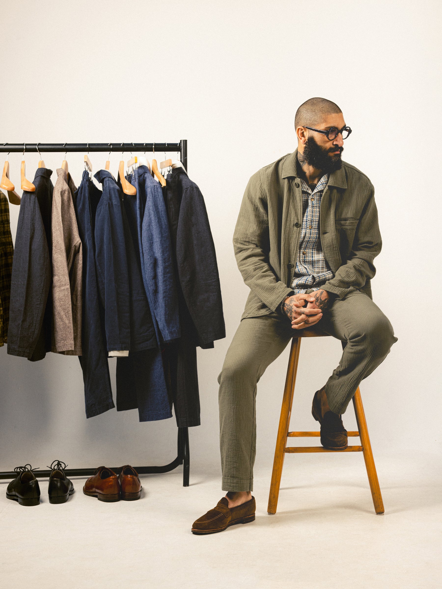 A man sat on a stool wearing an olive green suit, next to a clothing rail.