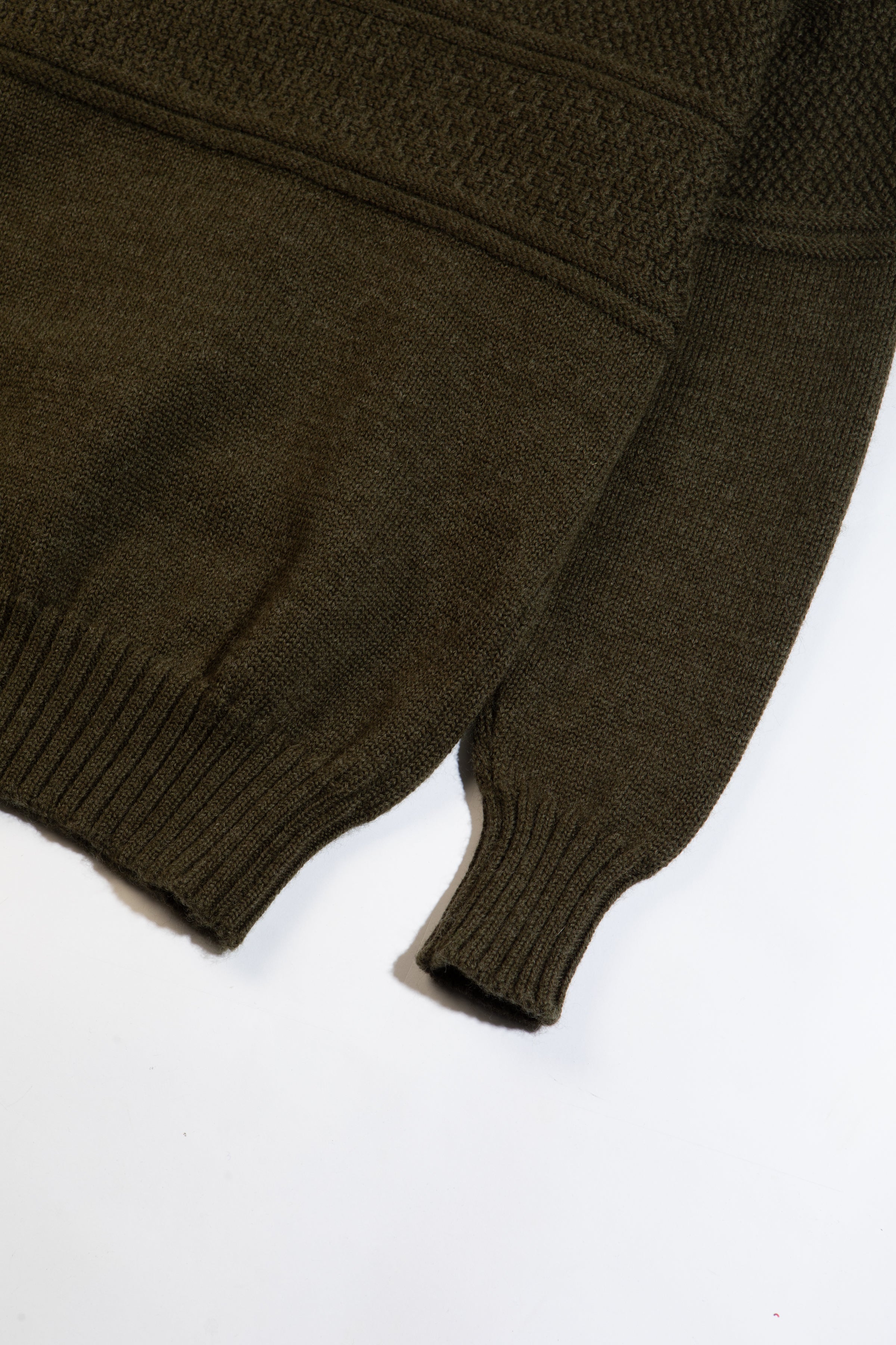 The ribbed hem and cuff of a knitted wool sweater in olive green, on a white background.