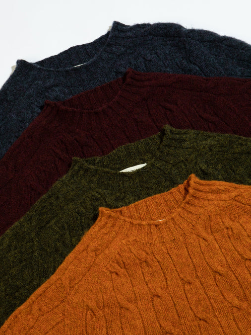 Four cable knit sweaters stacked on top of each other, in orange, red, green and blue.