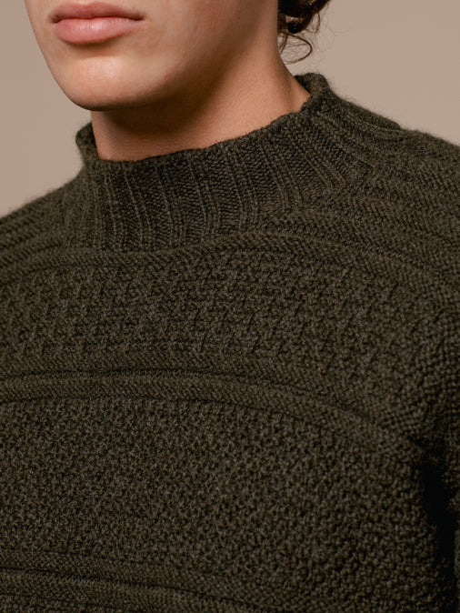 A man wearing a knitted sweater, made in Scotland in an olive green colour.