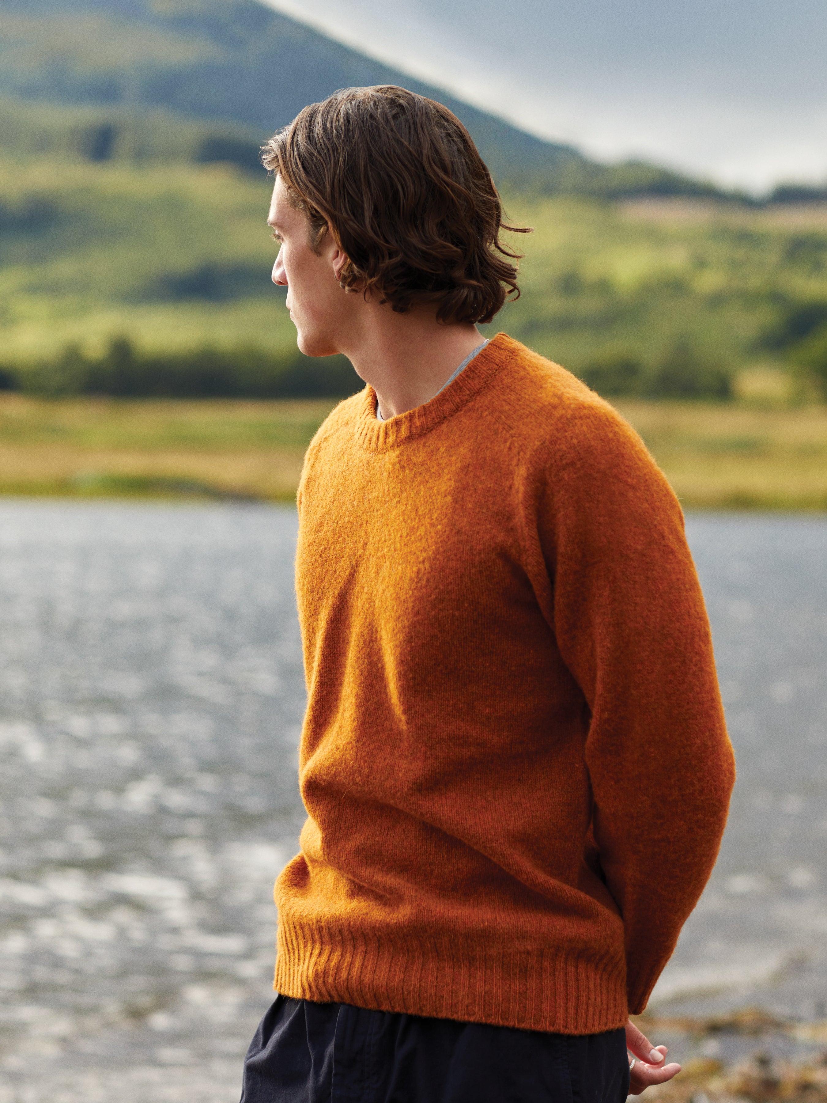 A man wearing a knitted crew neck sweater, made from Shetland wool in a tangerine orange.
