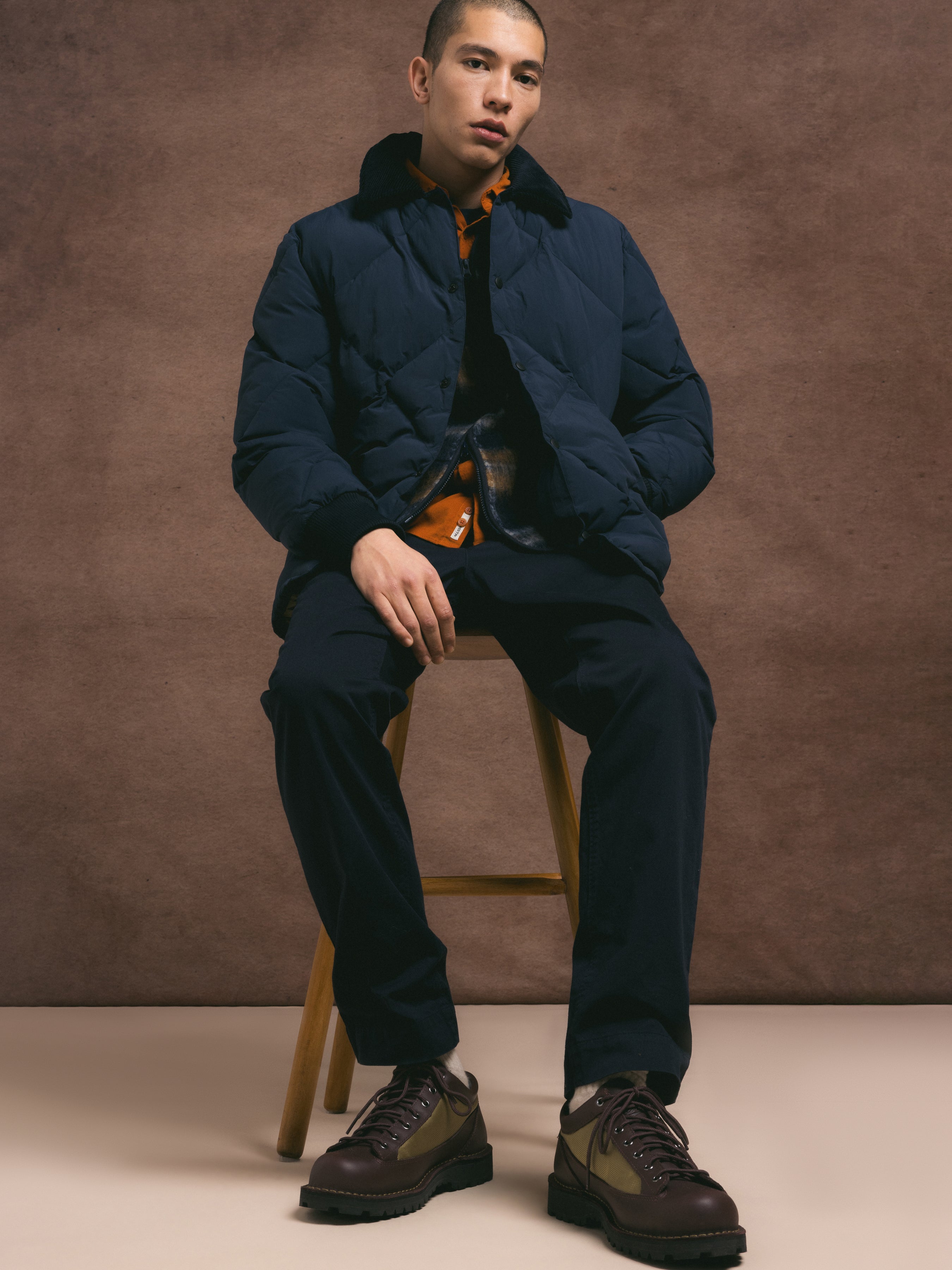 Man sitting on a stool wearing a dark blue puffer jacket and pants with brown shoes against a brown background