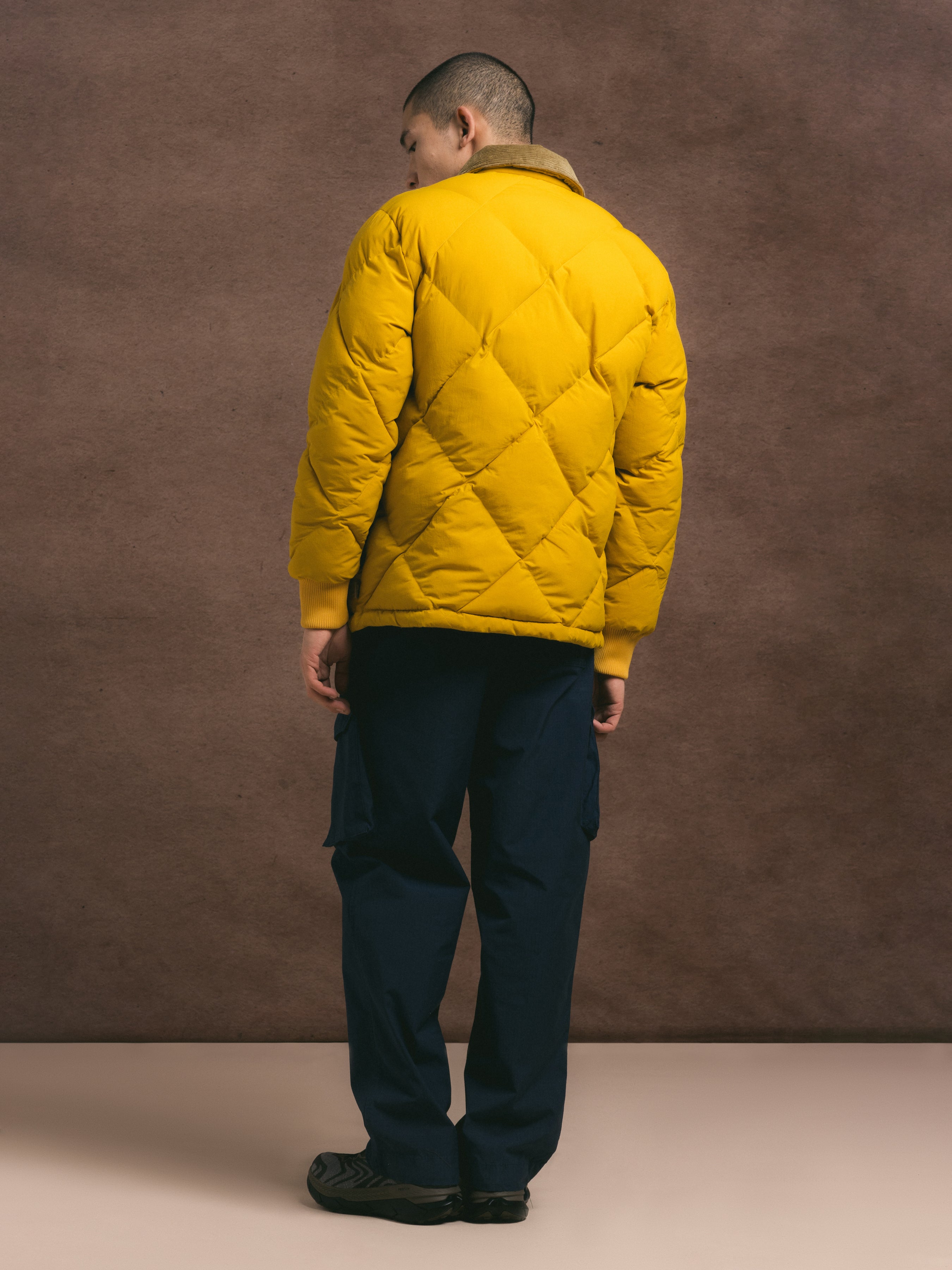 Person wearing a yellow quilted jacket and dark pants against a brown background