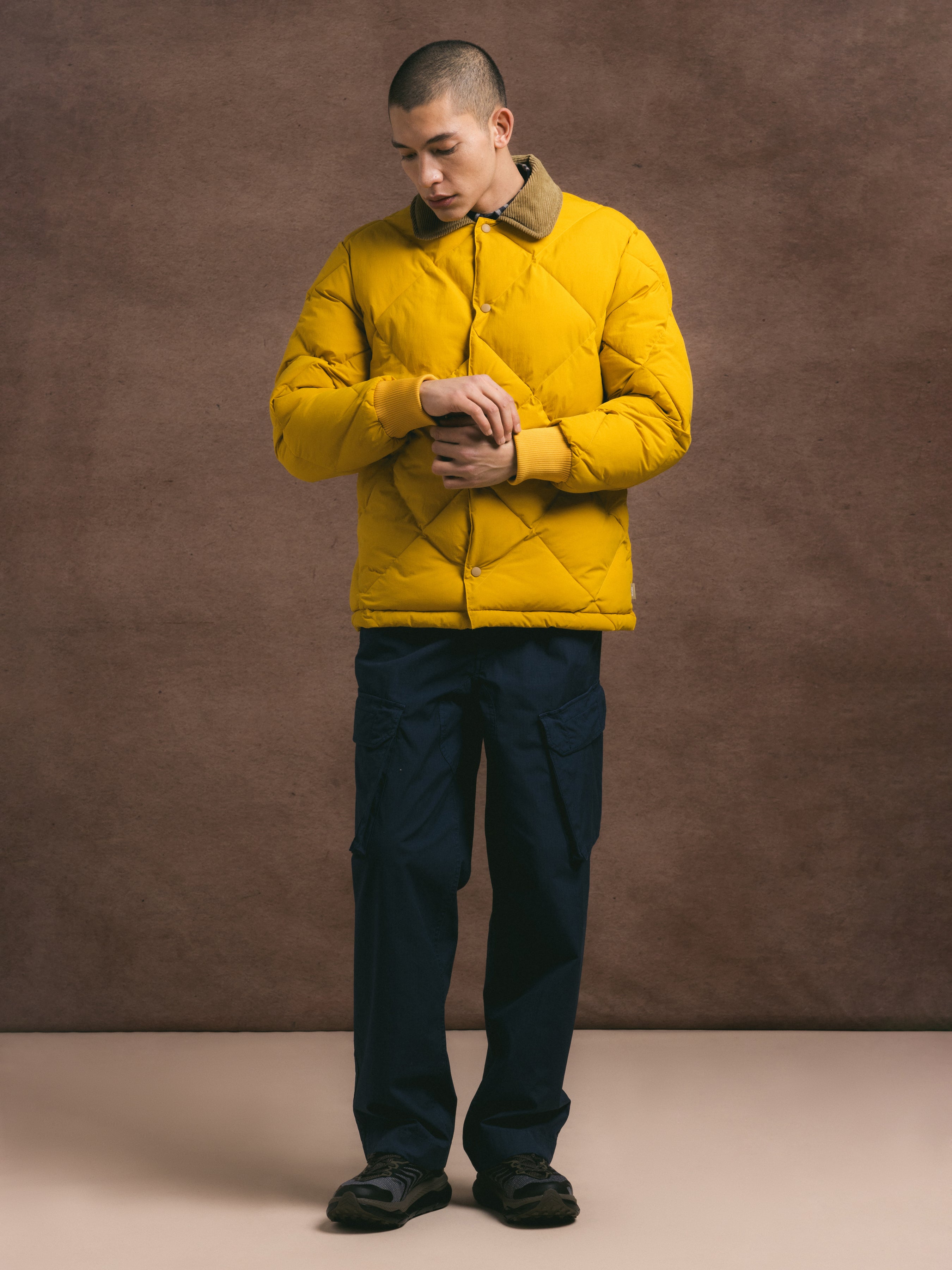 Man wearing a yellow puffer jacket and dark pants against a brown background