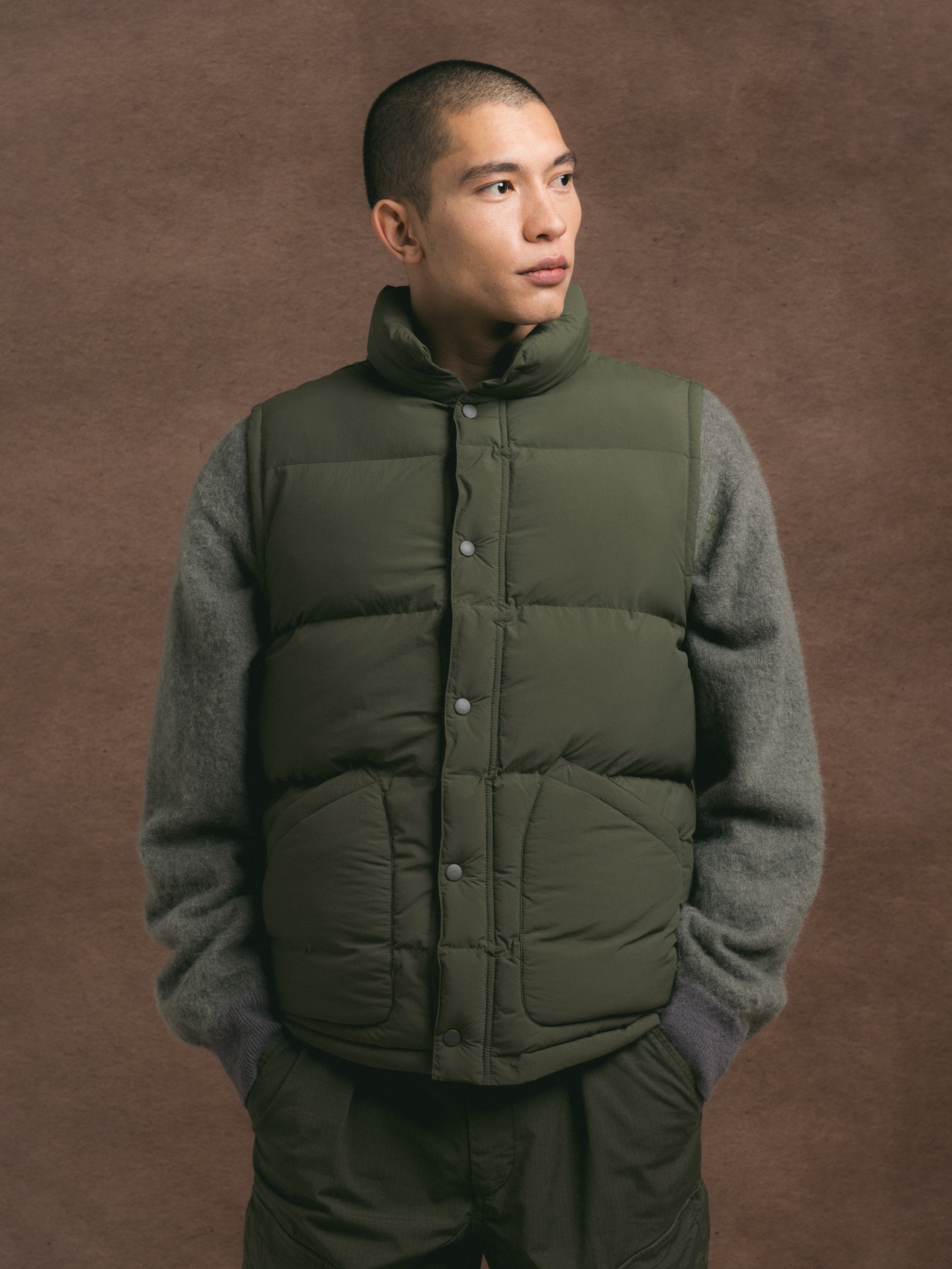 Man wearing a green puffer vest over a gray sweater against a brown background