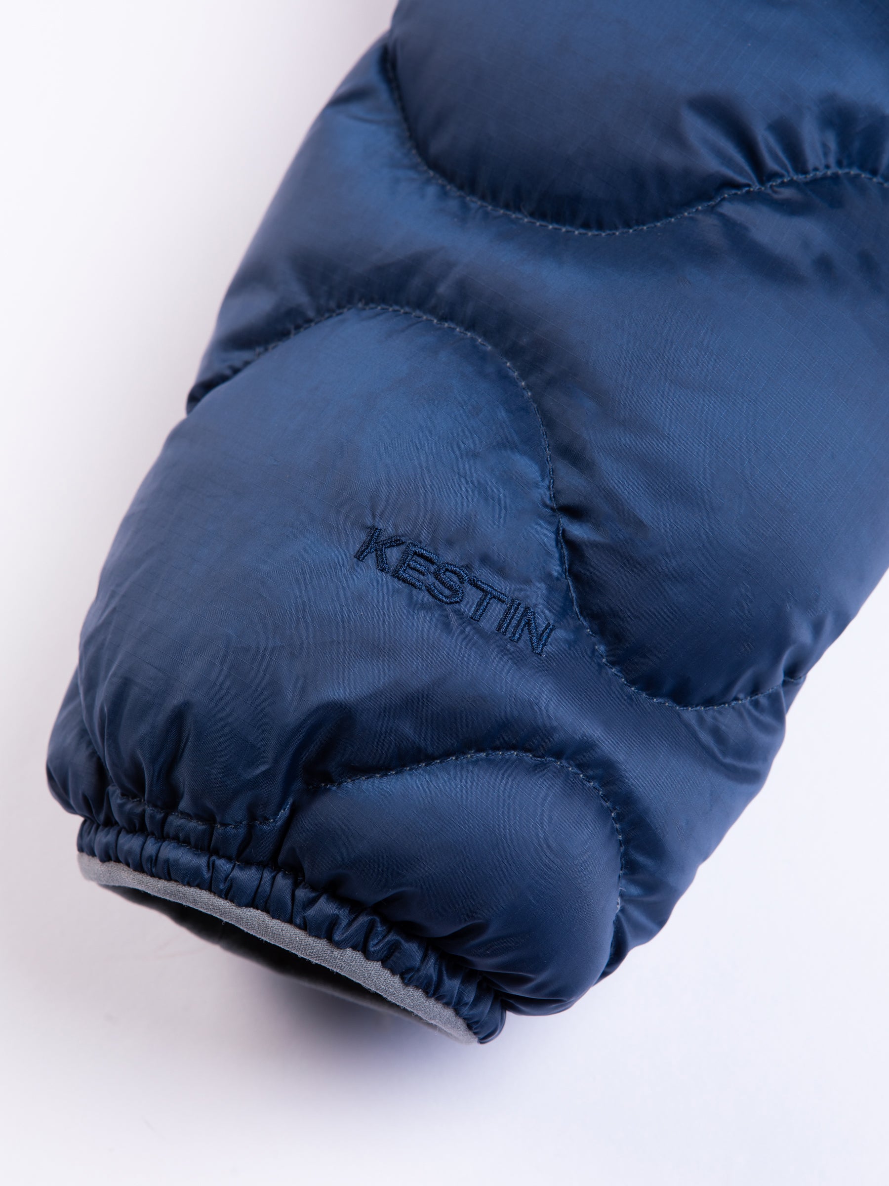 An embroidered KESTIN logo to the cuff of a blue quilted down jacket.