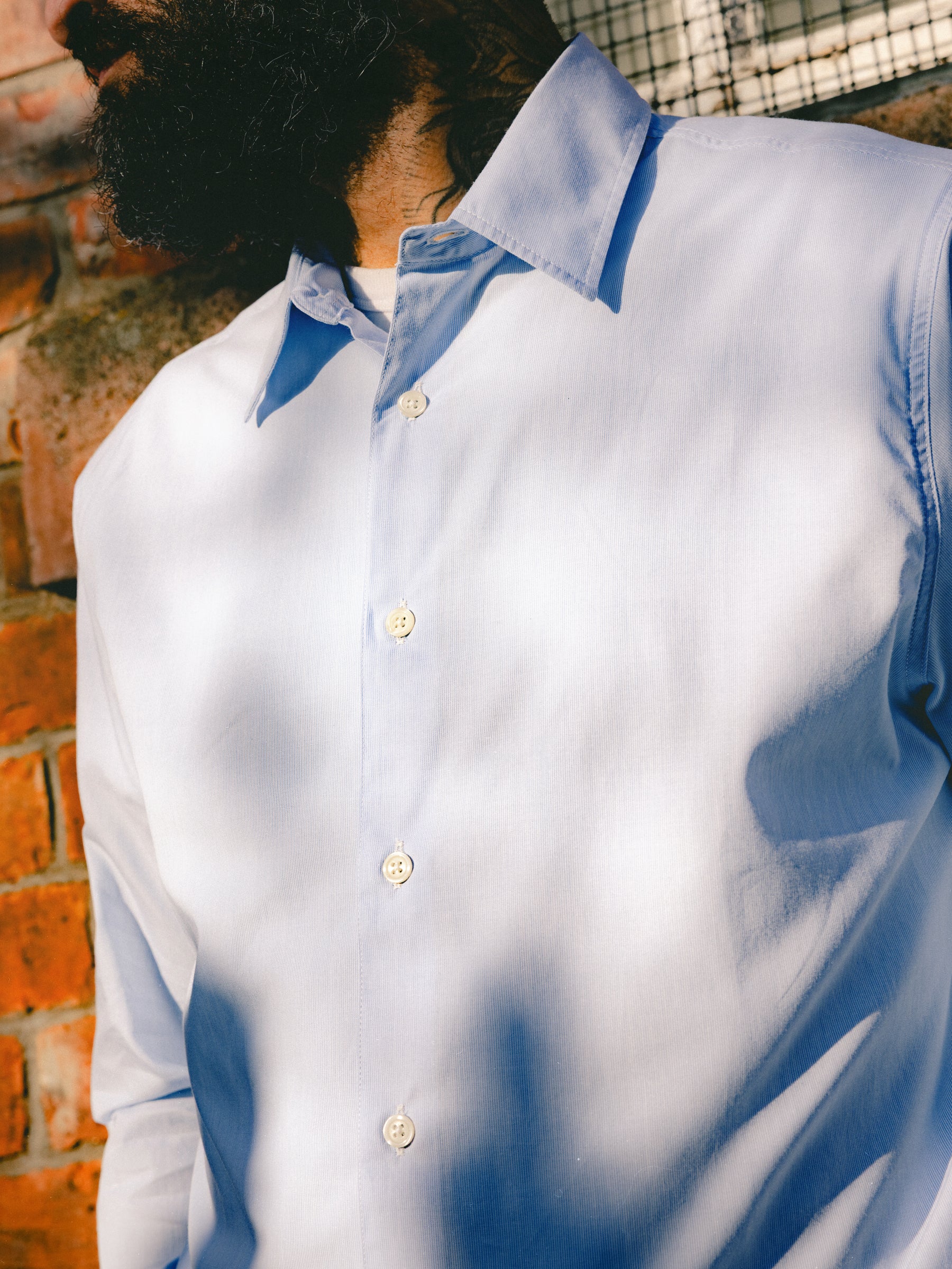 A man wearing a blue shirt with fine white stripes, standing againt a wall in the sun.