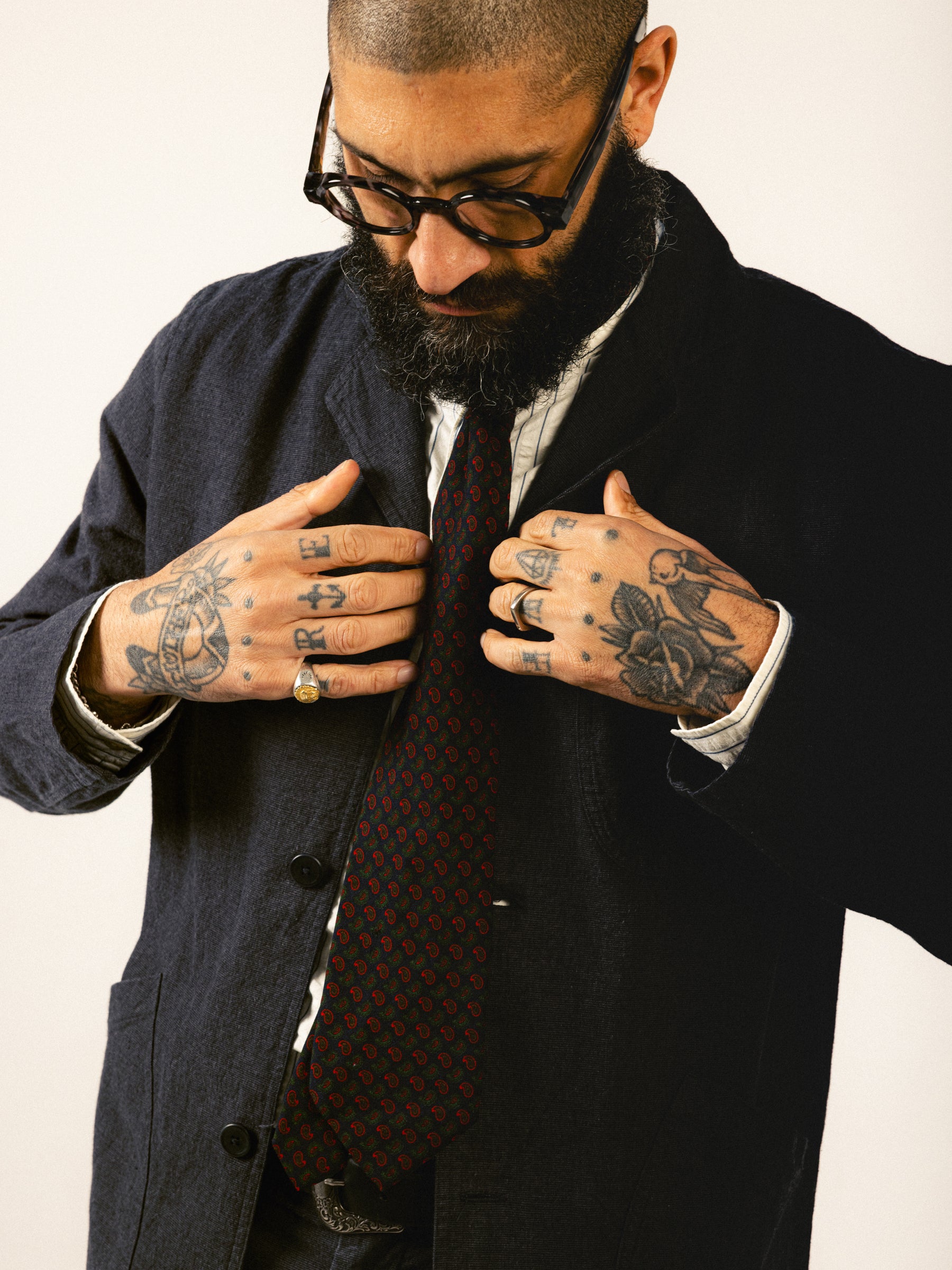 A man with glasses and traditional hand tattoos fixing the fit of his charcoal grey suit.