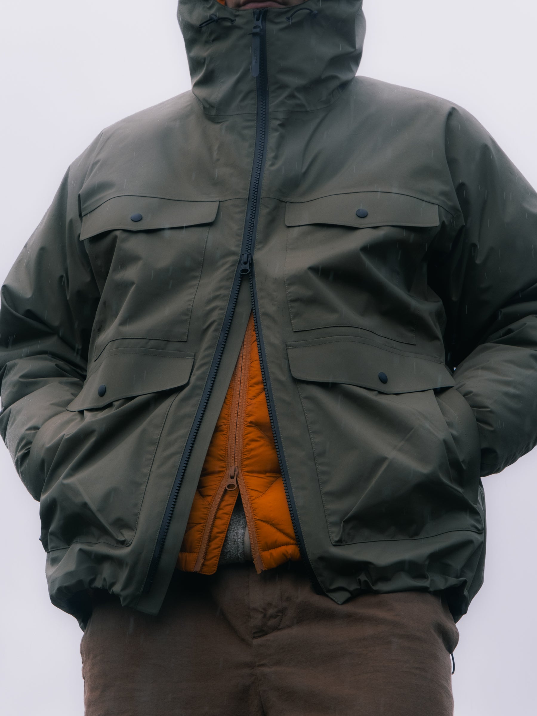 A man wearing a green waterproof jacket in the rain, layered over an orange down jacket.
