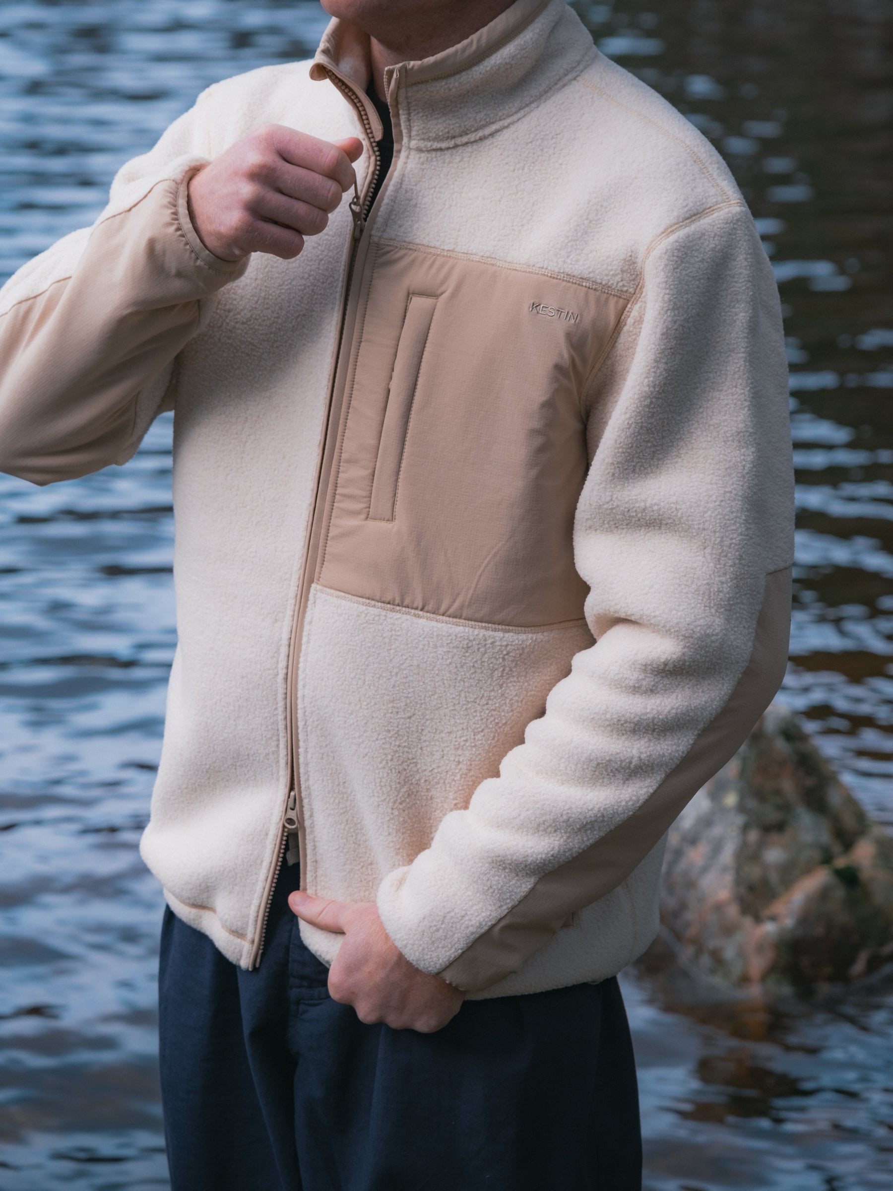 A man zipping up a cream fleece jacket with water in the background.