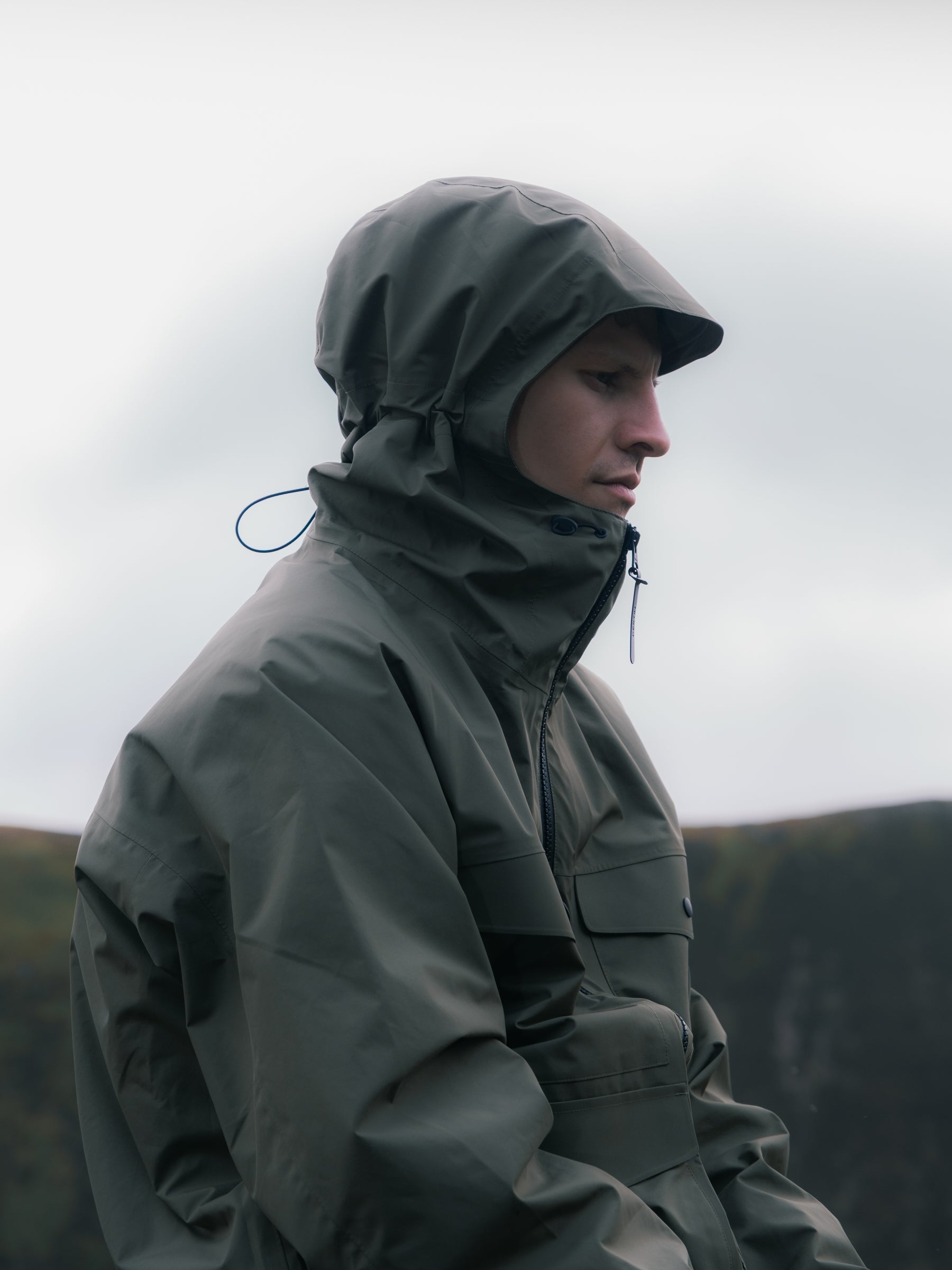 A man wearing a green waterproof jacket in the rain with his hood up.