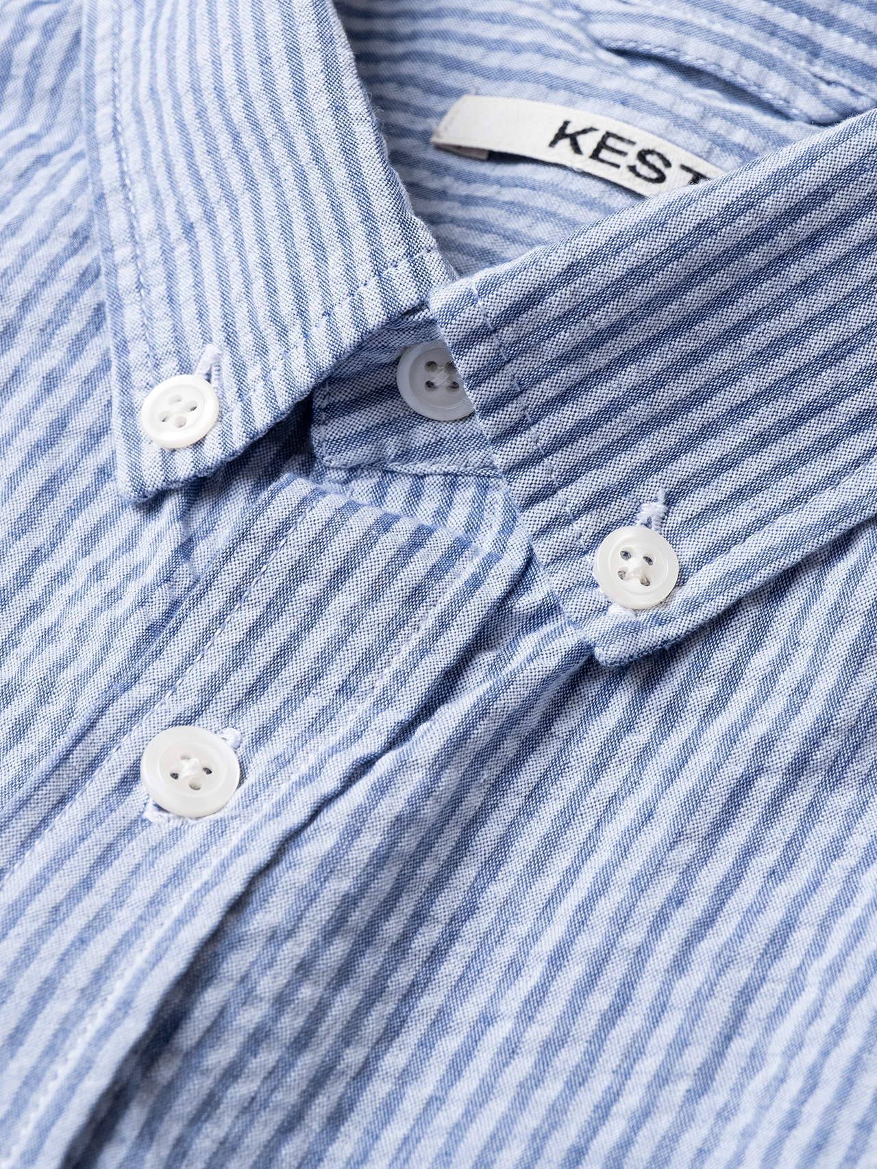 A close-up of a buttoned shirt collar from designer men's brand KESTIN, with a blue striped seersucker material.